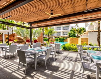 Outdoor patio with tables, a ceiling fan, and greenery, in a sunny, tropical resort setting.
