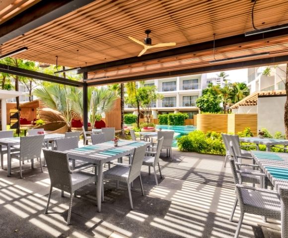 Outdoor patio with tables, a ceiling fan, and greenery, in a sunny, tropical resort setting.