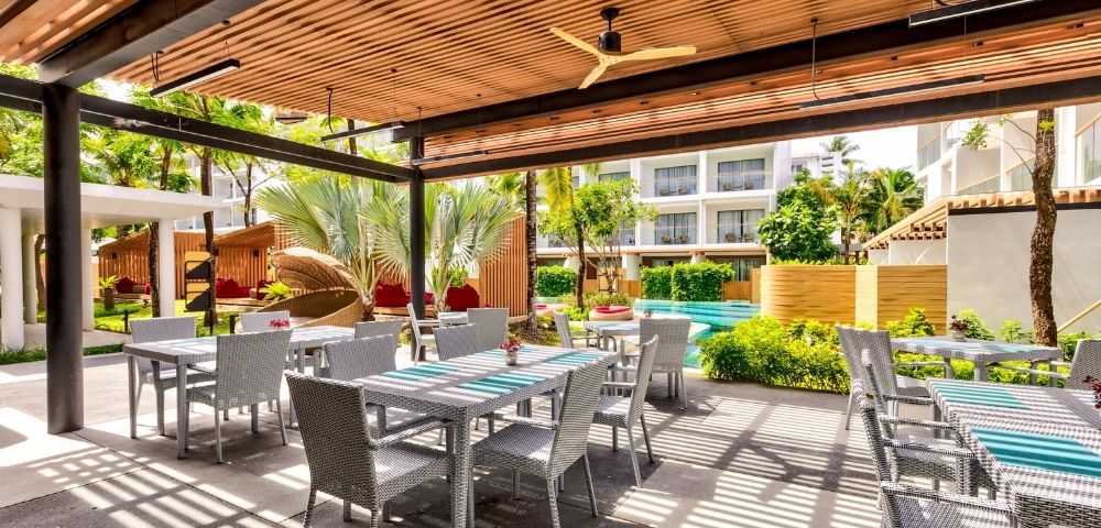 Outdoor patio with tables, a ceiling fan, and greenery, in a sunny, tropical resort setting.