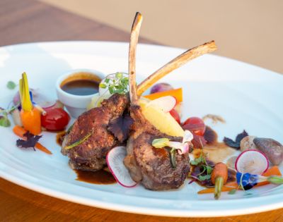 Lamb chops with herbs on a white plate, garnished with colorful vegetables and a small cup of sauce.