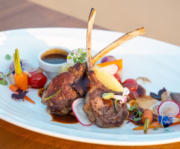 Lamb chops with herbs on a white plate, garnished with colorful vegetables and a small cup of sauce.