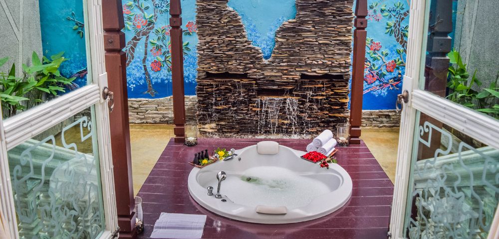 Tranquil spa retreat featuring a jacuzzi surrounded by a stone wall, colorful mural, and lush greenery, perfect for relaxation.