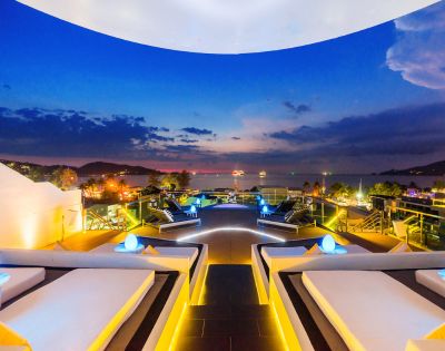 Luxurious rooftop lounge at dusk, featuring elegant seating, glowing lights, and a stunning ocean view against a colorful sky.