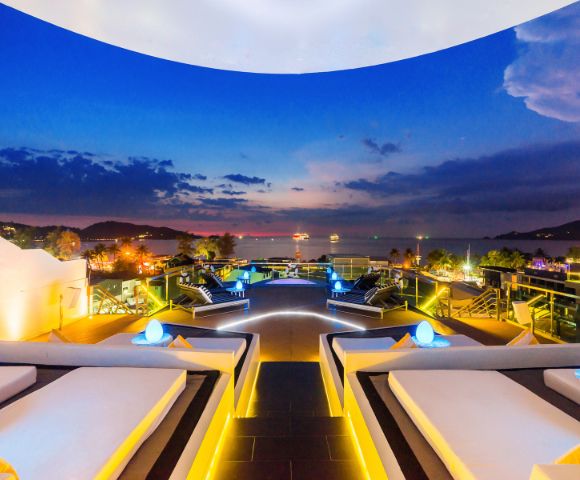 Luxurious rooftop lounge at dusk, featuring elegant seating, glowing lights, and a stunning ocean view against a colorful sky.