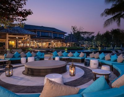 Outdoor lounge area with turquoise cushions, circular seating, lanterns, and modern architecture, set against a twilight sky.