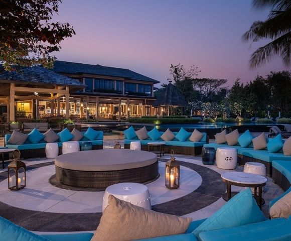 Outdoor lounge area with turquoise cushions, circular seating, lanterns, and modern architecture, set against a twilight sky.