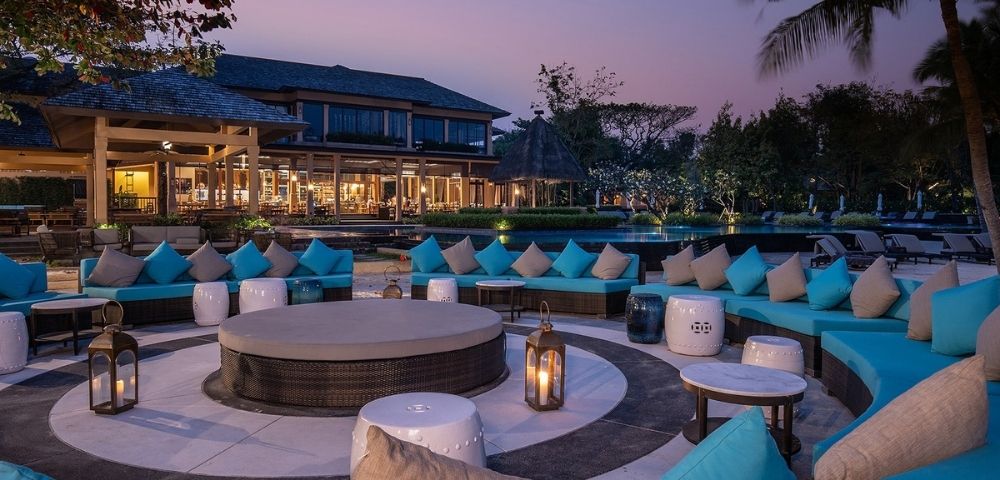 Outdoor lounge area with turquoise cushions, circular seating, lanterns, and modern architecture, set against a twilight sky.