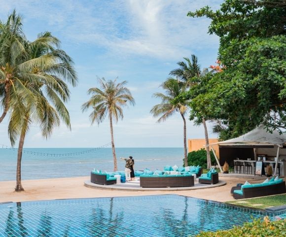 A tranquil beachside resort scene with palm trees, a blue pool, and outdoor seating facing the ocean, creating a serene atmosphere.