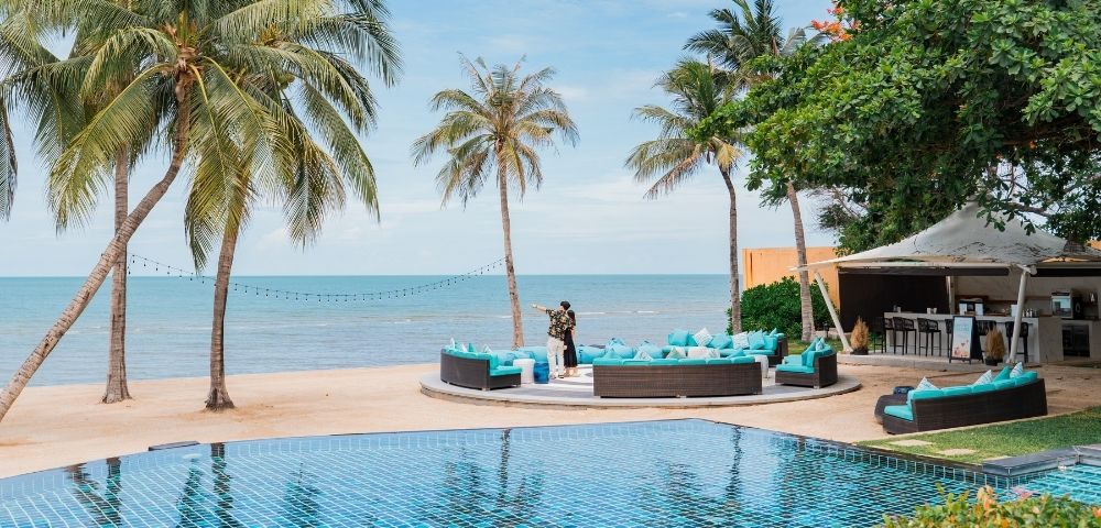 A tranquil beachside resort scene with palm trees, a blue pool, and outdoor seating facing the ocean, creating a serene atmosphere.