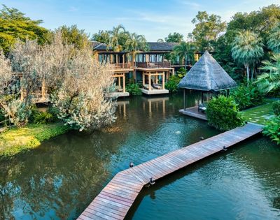 A serene resort featuring wooden pathways, lush greenery, and a thatched-roof gazebo beside a tranquil pond.