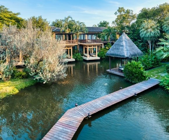 A serene resort featuring wooden pathways, lush greenery, and a thatched-roof gazebo beside a tranquil pond.