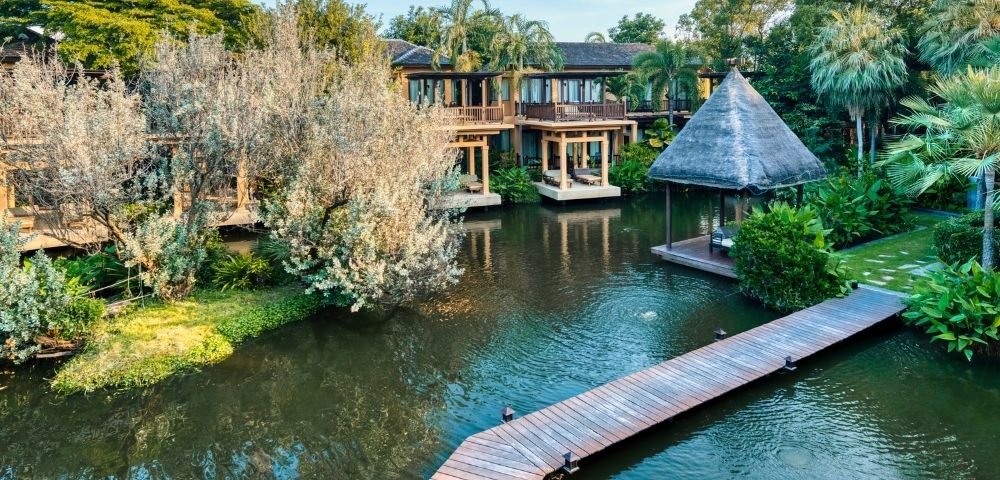 A serene resort featuring wooden pathways, lush greenery, and a thatched-roof gazebo beside a tranquil pond.