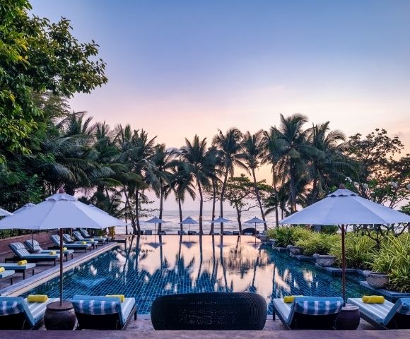 Serene resort pool surrounded by palm trees, lounge chairs, and umbrellas, with a beautiful sunset reflecting on the water.