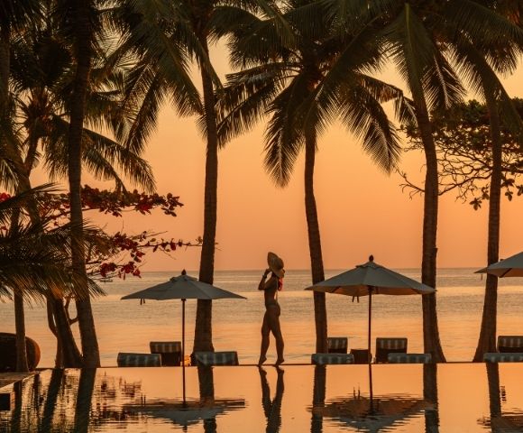Silhouetted woman in a hat walking by a tranquil pool surrounded by palm trees at sunset, creating a serene tropical atmosphere.