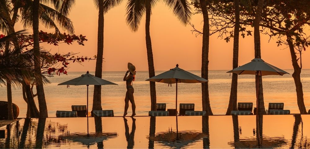 Silhouetted woman in a hat walking by a tranquil pool surrounded by palm trees at sunset, creating a serene tropical atmosphere.