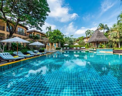 Resort pool glistening under a blue sky, surrounded by lush greenery, lounge chairs, and thatched-roof cabanas for relaxation.