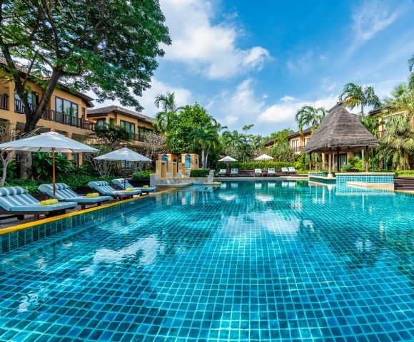 Resort pool glistening under a blue sky, surrounded by lush greenery, lounge chairs, and thatched-roof cabanas for relaxation.