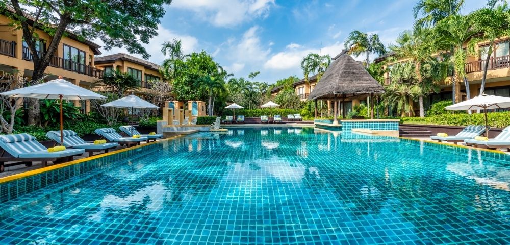 Resort pool glistening under a blue sky, surrounded by lush greenery, lounge chairs, and thatched-roof cabanas for relaxation.