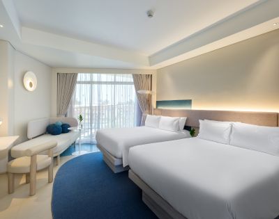 Bright hotel room featuring two queen beds, modern decor, a cozy seating area, and large windows with natural light.