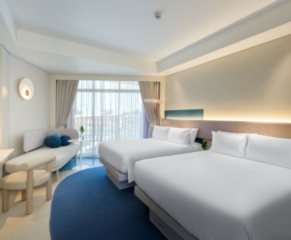 Bright hotel room featuring two queen beds, modern decor, a cozy seating area, and large windows with natural light.