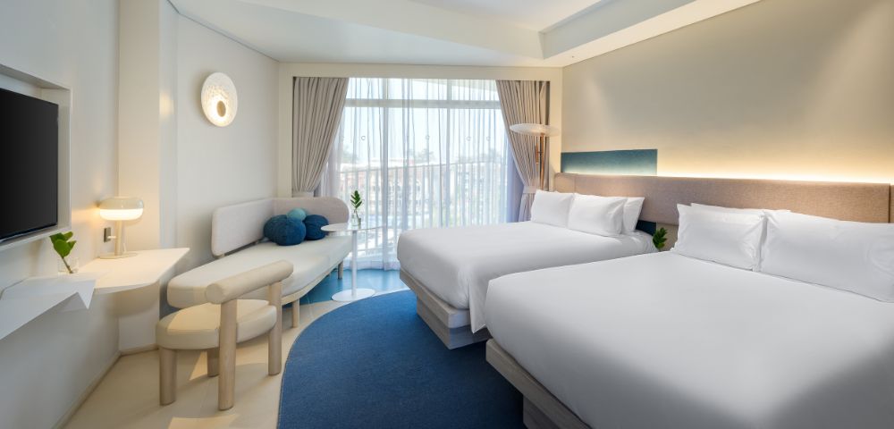 Bright hotel room featuring two queen beds, modern decor, a cozy seating area, and large windows with natural light.