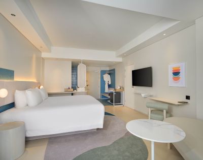 Modern hotel room featuring a large bed, stylish decor, a seating area, and a mounted TV against a light, airy backdrop.