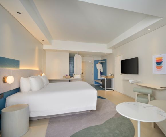 Modern hotel room featuring a large bed, stylish decor, a seating area, and a mounted TV against a light, airy backdrop.