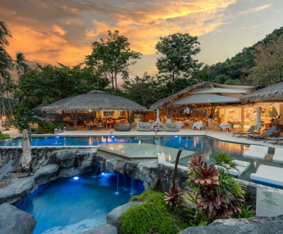 Tropical resort at sunset featuring a serene pool area, lush greenery, and thatched-roof lounges, creating a tranquil atmosphere.