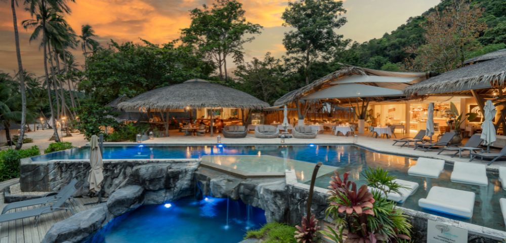 Tropical resort at sunset featuring a serene pool area, lush greenery, and thatched-roof lounges, creating a tranquil atmosphere.