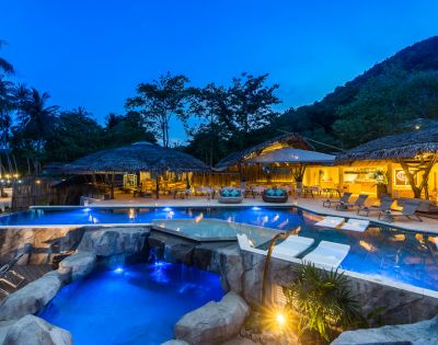 A serene resort with illuminated pools surrounded by tropical trees, featuring comfortable lounge areas and a cozy night ambiance.