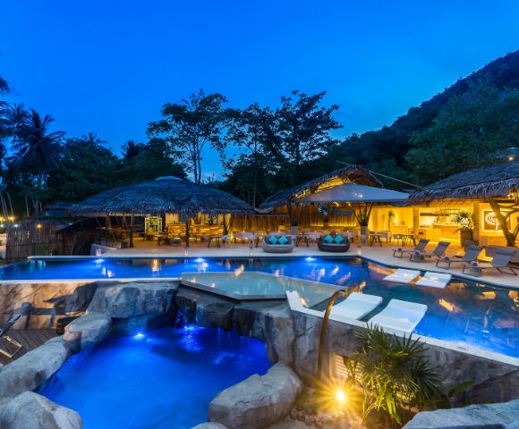 A serene resort with illuminated pools surrounded by tropical trees, featuring comfortable lounge areas and a cozy night ambiance.