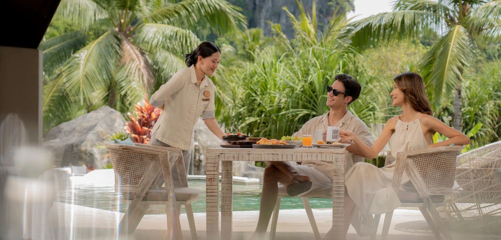 A serene outdoor dining scene with a waiter serving food to two guests amid lush tropical greenery and a sparkling pool.