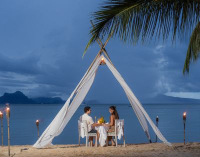 A romantic beach dinner setup under a fabric canopy, featuring a couple dining against a serene ocean backdrop at dusk.