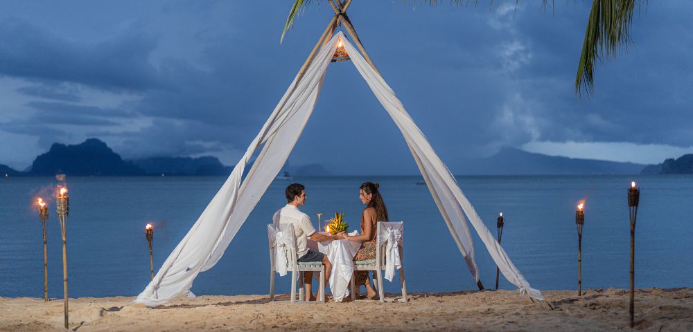 A romantic beach dinner setup under a fabric canopy, featuring a couple dining against a serene ocean backdrop at dusk.