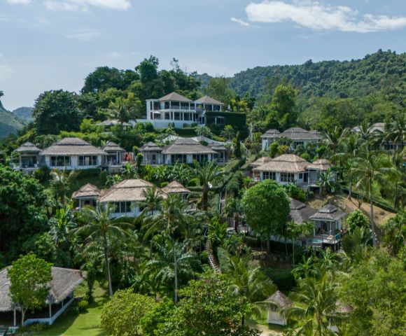 Aerial view of a lush hillside resort featuring several tropical villas surrounded by greenery and palm trees under a blue sky.