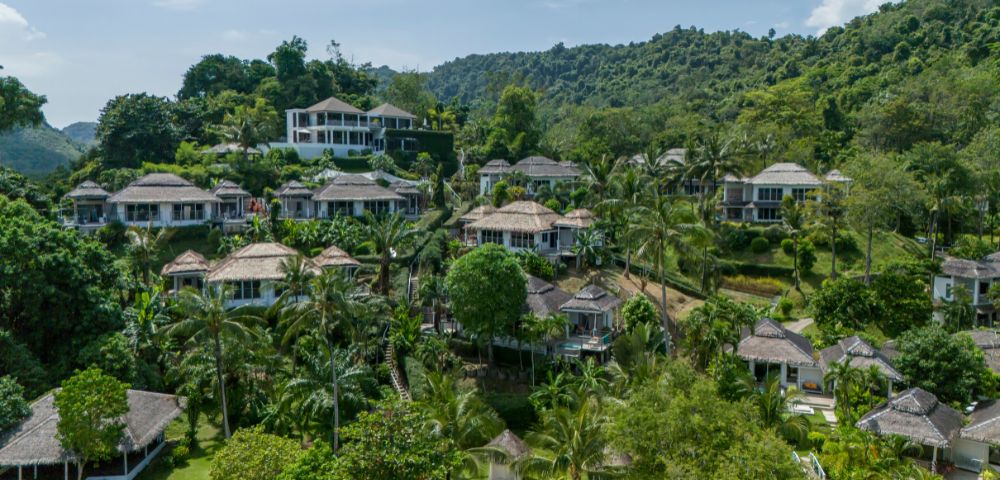 Aerial view of a lush hillside resort featuring several tropical villas surrounded by greenery and palm trees under a blue sky.