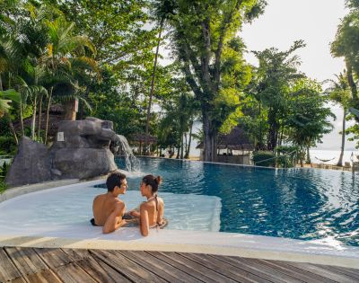 A couple relaxes in a serene pool surrounded by lush greenery and a waterfall, enjoying a tranquil tropical setting.