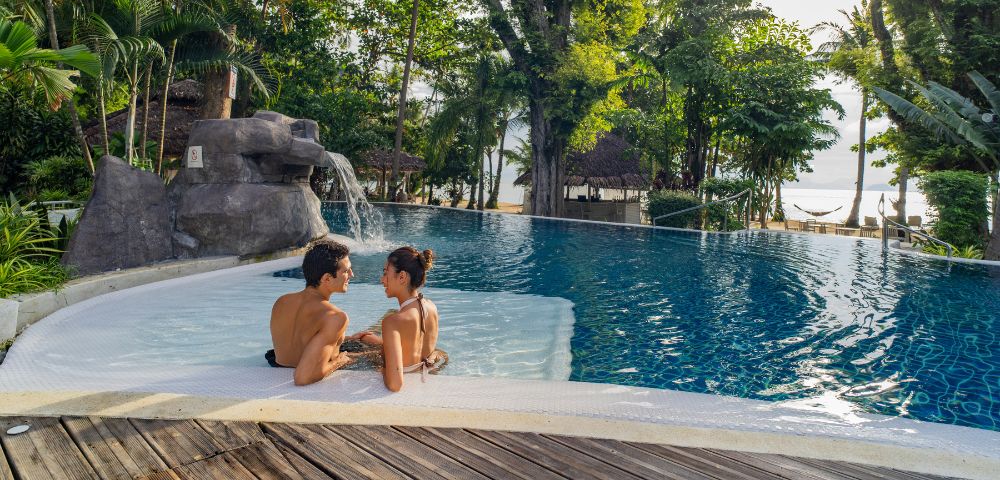 A couple relaxes in a serene pool surrounded by lush greenery and a waterfall, enjoying a tranquil tropical setting.
