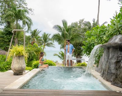 A couple enjoys a private pool surrounded by lush greenery and palm trees, with a serene ocean view in the background.