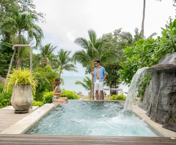A couple enjoys a private pool surrounded by lush greenery and palm trees, with a serene ocean view in the background.