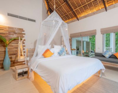 A serene hotel room featuring a canopied bed, vibrant throw pillows, wooden accents, and large windows with lush greenery outside.