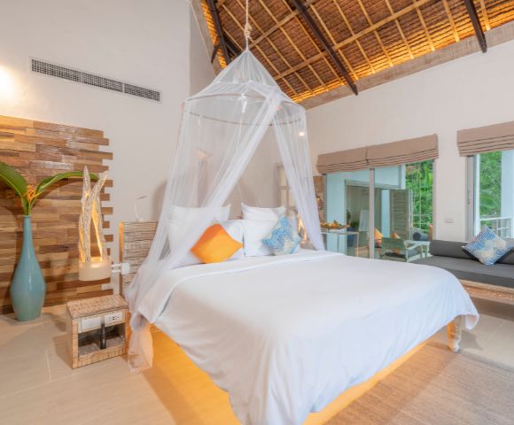 A serene hotel room featuring a canopied bed, vibrant throw pillows, wooden accents, and large windows with lush greenery outside.
