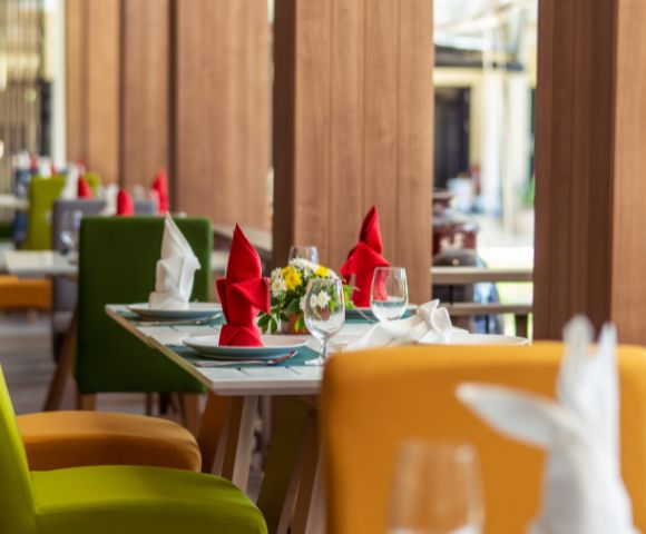 Colorful dining area with green, yellow, and red chairs; elegantly set tables with glassware, napkins, and floral centerpieces.
