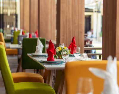 Colorful dining area with green, yellow, and red chairs; elegantly set tables with glassware, napkins, and floral centerpieces.