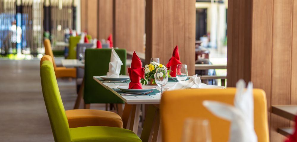 Colorful dining area with green, yellow, and red chairs; elegantly set tables with glassware, napkins, and floral centerpieces.