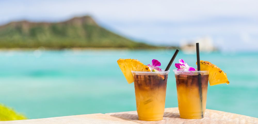 Two tropical drinks with pineapple and orchid garnishes sit on a table, overlooking a serene turquoise ocean and distant mountains.