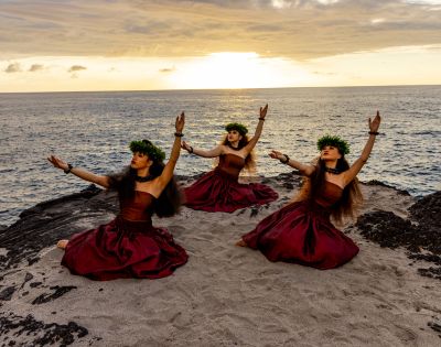 Three hula dancers in red dresses and flower crowns perform gracefully on a sandy beach at sunset, framed by a tranquil ocean.