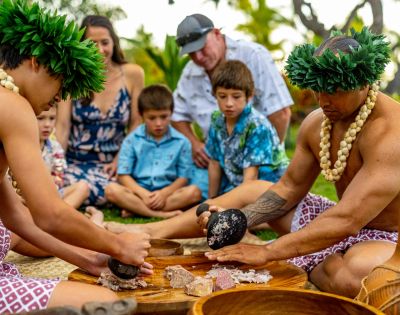 Two individuals in traditional attire prepare food using stone tools, surrounded by a group of attentive children in a lush outdoor setting.