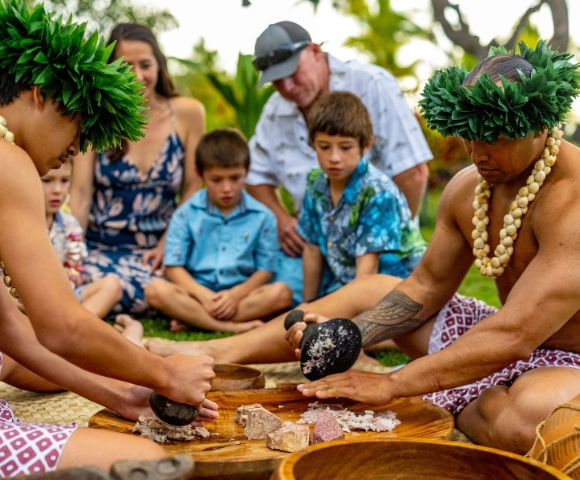 Two individuals in traditional attire prepare food using stone tools, surrounded by a group of attentive children in a lush outdoor setting.