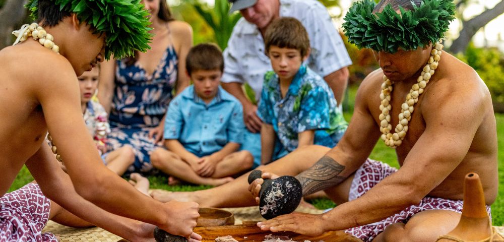 Two individuals in traditional attire prepare food using stone tools, surrounded by a group of attentive children in a lush outdoor setting.